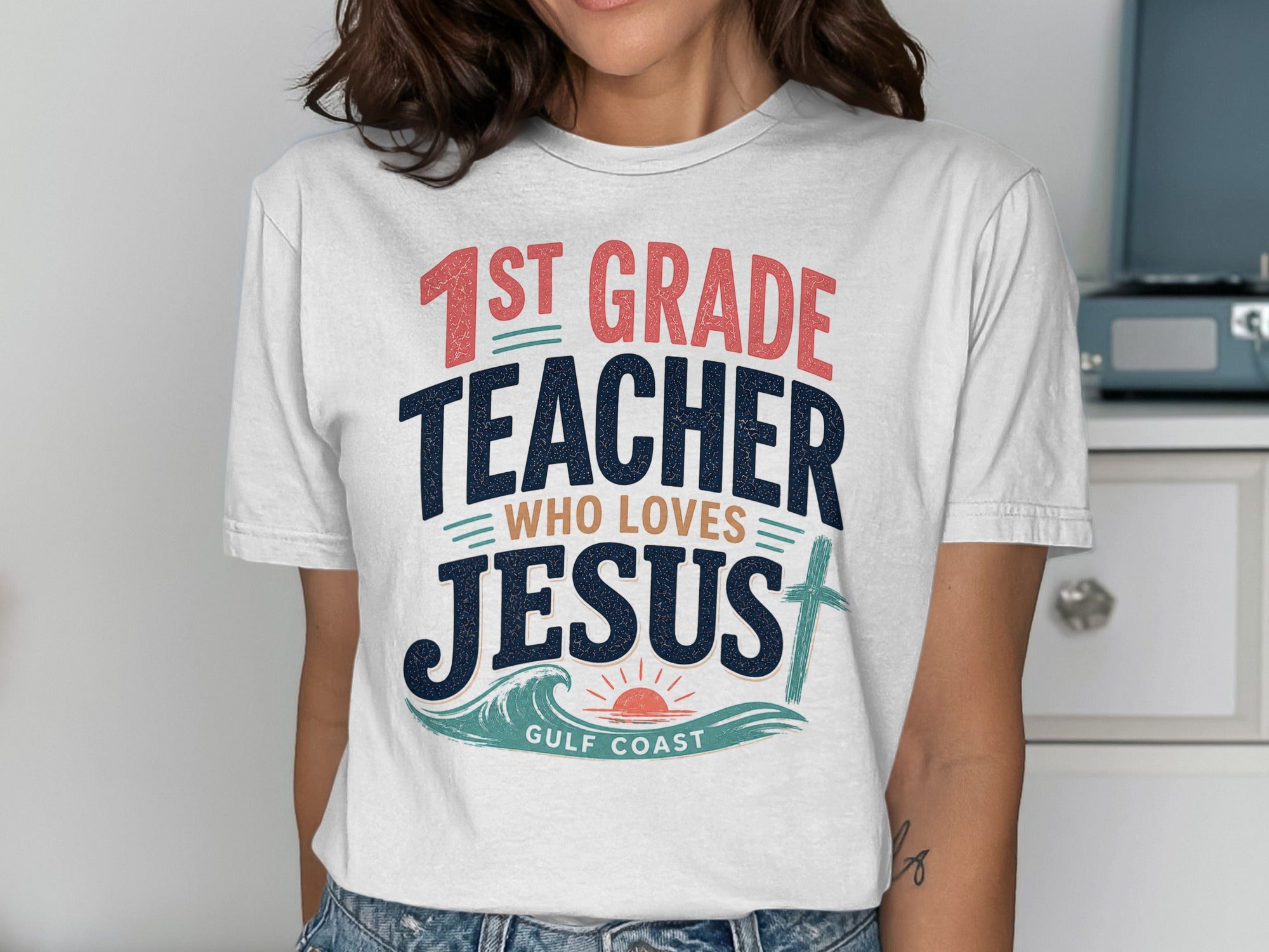 White t-shirt with "1st Grade Teacher Who Loves Jesus" text and ocean graphic.