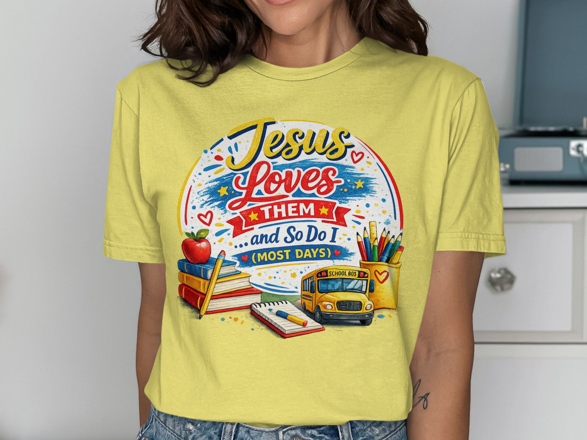 Yellow t-shirt with "Jesus Loves Them (Most Days)" text, school bus, and art supplies.