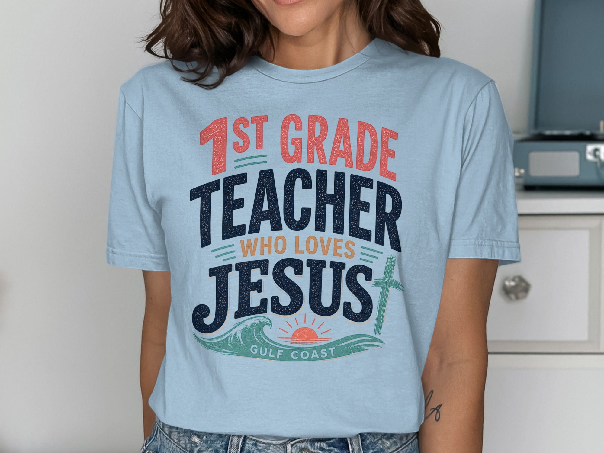 Light blue t-shirt with "1st Grade Teacher Who Loves Jesus" text and ocean waves.