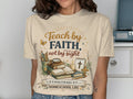 Cream t-shirt with "Teach by Faith, Not by Sight" and homeschool theme graphic.